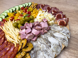 Charcuterie board with cucumber, chorizo, smoked ham, figs, canned ham, head cheese, cheese cubes, tomatoes, olives, breadsticks and crackers on wooden table, party appetizer.