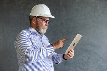 An older engineer wearing a white safety helmet uses a digital tablet, symbolizing modern construction practices, technology integration, and innovation in engineering.
