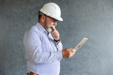 An older engineer wearing a white safety helmet uses a digital tablet, symbolizing modern construction practices, technology integration, and innovation in engineering.