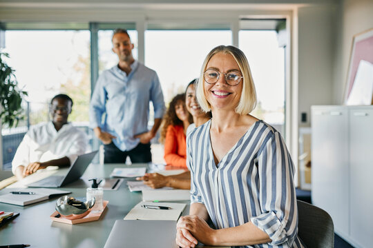 A group of diverse colleagues look into the camera smiling. They are sitting in a new, stylish office, with notebooks, paper and a laptop on the table