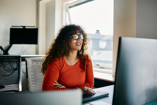 Young Professional Woman Working Smiling in Modern Office Environment. She is wearing an orange sweater, beautiful curly hair and black glasses. She is looking intently at a screen.