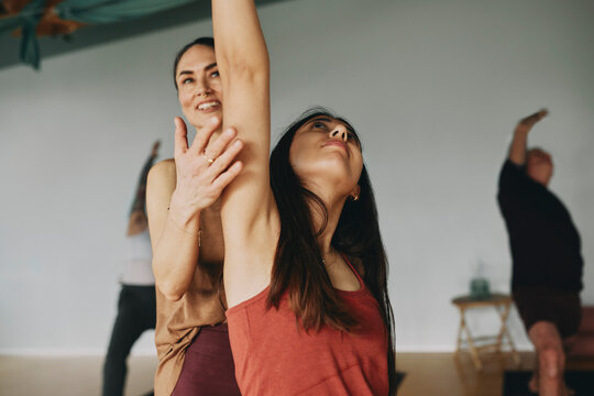 Young female yoga instructor smiling while helping a young woman with her stretching during a class in her studio