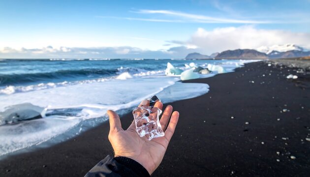 A clear ice shard held in a hand, showcasing a dramatic black sand beach and ocean waves, bathed in vibrant sunlight.