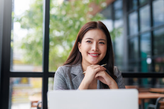 Smiling Asian businesswoman working on a laptop in a stylish cafe, enjoying a productive moment filled with natural light and greenery