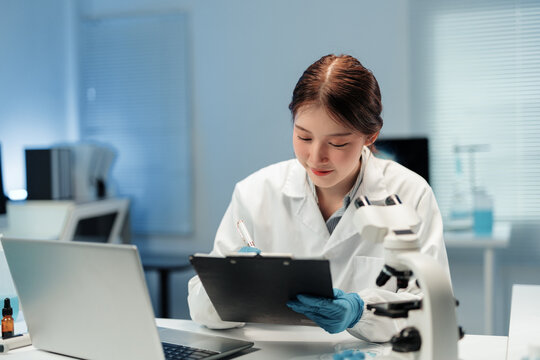 Asian female scientist wearing lab coat and gloves taking notes on clipboard while using microscope and laptop in modern laboratory