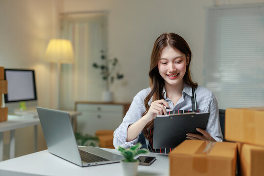 Smiling young entrepreneur checking notes on clipboard while using laptop for online sales and order management in her home office