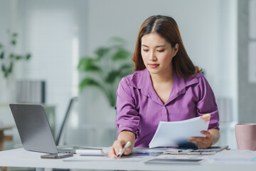 Young Asian businesswoman analyzing documents while working on a laptop in a modern office, surrounded by a bright, organized workspace