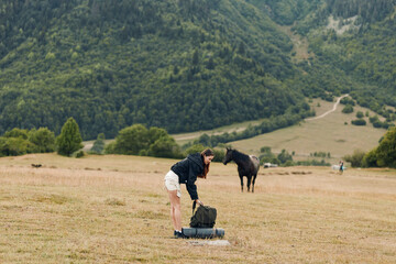 A person wearing a dark jacket and light shorts bends to adjust a backpack on a sunlit field, with a horse grazing nearby and distant hills under a clear sky.