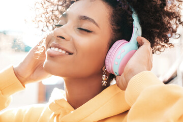 Beautiful woman with afro curls hairstyle.Smiling model in yellow hoodie.Sexy carefree female enjoying listening music in wireless headphones.Posing on street background at sunset.Closed eyes