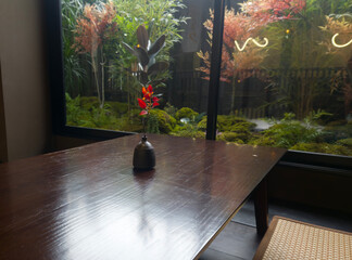 Serene Garden View: A tranquil scene unfolds, as a minimalist setting inside gently complements the vibrant garden outside. The focus is drawn to a sleek wooden table, on which rests a single.
