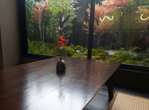 Serene Garden View: A tranquil scene unfolds, as a minimalist setting inside gently complements the vibrant garden outside. The focus is drawn to a sleek wooden table, on which rests a single.