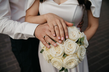 A bride in a white dress and a groom in a white shirt on their wedding day placed their palms on top of each other, showing off the gold wedding rings on their fingers, above a bouquet of white roses