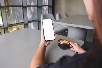 Mockup image of a woman holding and using mobile phone with blank desktop screen while drinking coffee in cafe