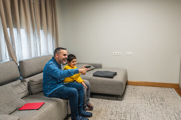 Happy father and son enjoying time together, sitting on sofa and watching television, pointing at screen with remote control in hand
