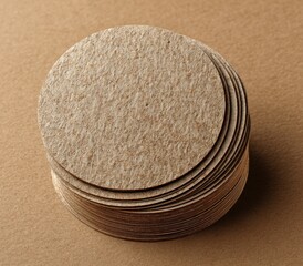 A neatly stacked pile of round, light brown, textured cardboard coasters on a matching surface