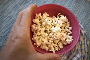 Close-up of popcorn in a pastel pink bowl placed on a blue checkered kitchen towel. Minimal composition with copy space and hand.