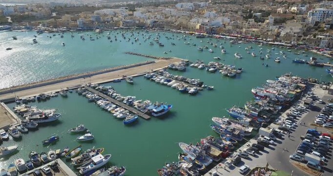Malta: Aerial view of Marsaxlokk harbor in Malta, featuring numerous colorful traditional Luzzu fishing boats and yachts moored in the turquoise waters on a summer sunny day. Drone flight footage