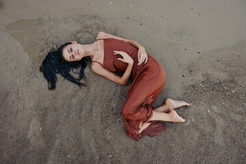 Woman lying on sandy beach in a flowing rust-colored dress, with relaxed and dreamy expression in a natural outdoor setting.