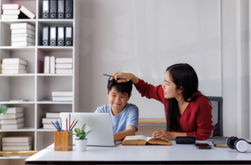 Asian mother helping son with homeschooling on laptop, pointing at screen with pencil