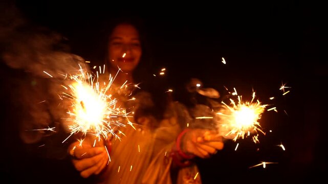 Happy Indian woman celebrating Diwali festival with holding sparklers in hand. Lady wearing a traditional saree. Enjoying with fire crackers in dark night background.