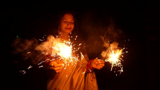 Happy Indian woman celebrating Diwali festival with holding sparklers in hand. Lady wearing a traditional saree. Enjoying with fire crackers in dark night background.