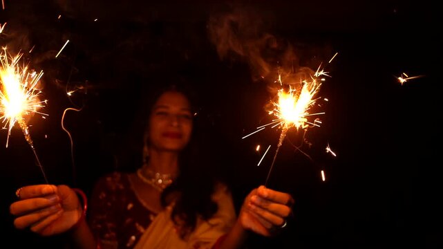 Happy Indian woman celebrating Diwali festival with holding sparklers in hand. Lady wearing a traditional saree. Enjoying with fire crackers in dark night background.