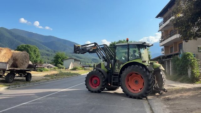 A green agricultural tractor uses its front loader to move large round hay bales on a sunny day. The scene is set in a picturesque rural area with rolling green mountains and clear blue skies.