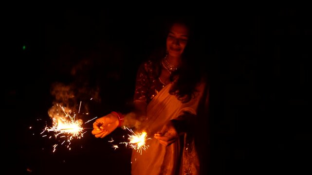 Happy Indian woman celebrating Diwali festival with holding sparklers in hand. Lady wearing a traditional saree. Enjoying with fire crackers in dark night background.