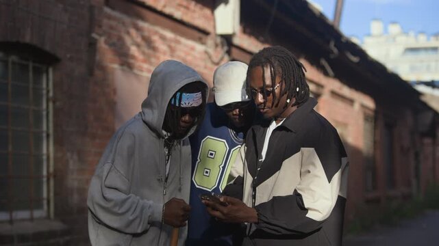 Group of young Black men wearing y2k hip-hop outfits standing outdoors looking at phone screen and talking, whip pan shot