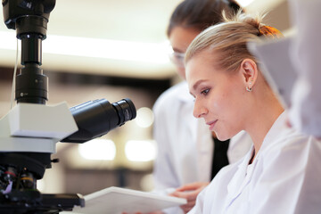 A team of female professionals works together in a science lab. One woman looks into a microscope while her colleagues help and discuss the results of their project.
