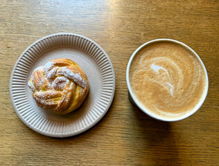 Coffee latte art on wood table with a bun with lemon in cafe close-up. Cappuccino with a pattern in a paper cup and bun on a wooden table in a coffee shop top view. The concept of food and drinks.