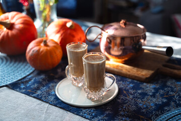 Two glass mugs of creamy pumpkin spice latte topped with cinnamon on a white plate. Autumn coffee drink served on a cozy festive table decorated with pumpkins, copper pot, and rustic details. 