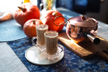 Two glass mugs of creamy pumpkin spice latte topped with cinnamon on a white plate. Autumn coffee drink served on a cozy festive table decorated with pumpkins, copper pot, and rustic details. 