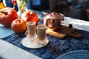 Two glass mugs of creamy pumpkin spice latte topped with cinnamon on a white plate. Autumn coffee drink served on a cozy festive table decorated with pumpkins, copper pot, and rustic details. 