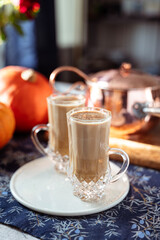 Two glass mugs of creamy pumpkin spice latte topped with cinnamon on a white plate. Autumn coffee drink served on a cozy festive table decorated with pumpkins, copper pot, and rustic details. 