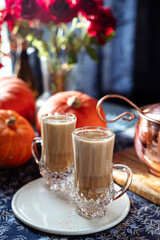 Two glass mugs of creamy pumpkin spice latte topped with cinnamon on a white plate. Autumn coffee drink served on a cozy festive table decorated with pumpkins, copper pot, and rustic details. 