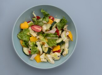 Bowl of mixed salad with lettuce leaves, cucumbers, cherry tomatoes, and mango. Healthy vegetarian food rich in vitamins, fresh organic meal for diet and wellness, isolated on gray background.