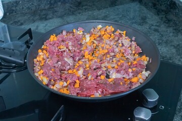 Close-up of raw beef with chopped vegetables, including onion and carrot, cooking in a frying pan on a modern kitchen stove. Fresh home-cooked food preparation concept.
