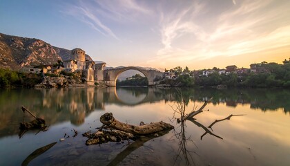 Fototapeta premium A serene sunrise view of the historic bridge, reflected perfectly in the tranquil water, with weathered driftwood along the shore.