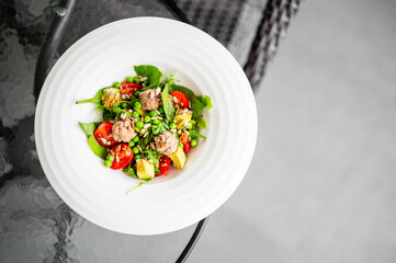 Fresh tuna salad with avocado, cherry tomatoes, green peas, and leafy greens served on a white plate. Healthy meal on glass table in natural light. Top view, indoor setting