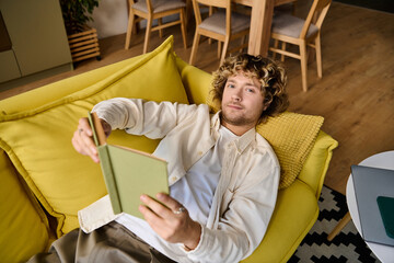 Handsome man lounging on a yellow sofa while reading a book during a relaxing afternoon