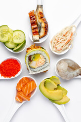 Sushi roll on a white background, and around it are white spoons with ingredients. Salmon, shrimp, tuna