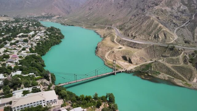 A scenic aerial view of the Naryn River flowing between a village and a winding mountain road, connected by a bridge.
