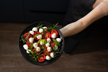 A person holds a black plate filled with vibrant slices of tomatoes, small mozzarella balls, and...