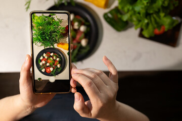 A person holds a smartphone, taking a picture of a colorful salad made with fresh vegetables like tomatoes and greens. The kitchen has a clean, modern design and various ingredients nearby.