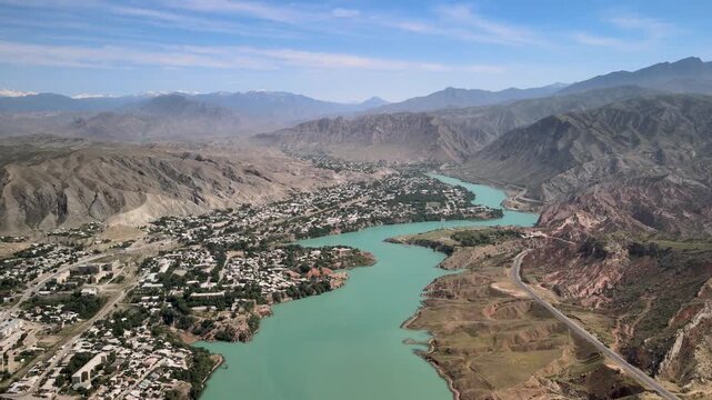 A scenic aerial view of the Naryn River flowing between a village and a winding mountain road.The river&rsquo;s vibrant turquoise waters contrast beautifully with the rugged mountain landscape of Kyrgyzstan