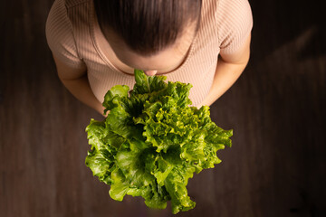 A woman stands indoors holding a large bunch of fresh green lettuce. She looks down at the lettuce...