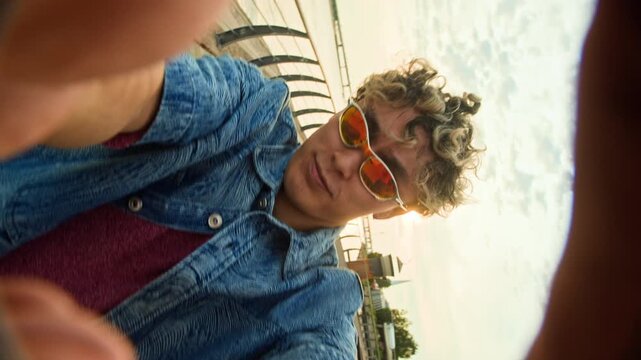 Medium POV shot of gen Z Asian guy in sunglasses gesturing and having fun on camera while walking on city embankment in summer, UGC