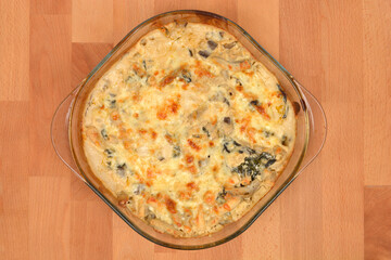A close-up top-down view of a glass dish filled with baked pasta, cheese, and chard, with a golden-brown melted cheese crust on a wooden background with copy space