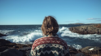 Individual with wavy hair wearing a patterned sweater stands on rocky shore, gazing at ocean waves crashing against the coastline under a clear blue sky, reflecting tranquility and nature